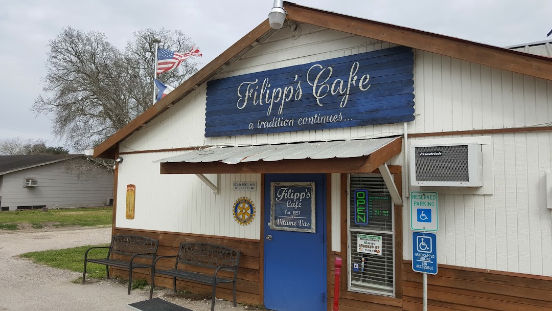 A small, single-story cafe with a blue sign reading "Filipp's Cafe" above the entrance. Flags fly on the roof, a wooden bench sits outside Filipp's Cafe, and handicap parking signs are visible near the door. - Market Design Team: Define. Structure. Expand.