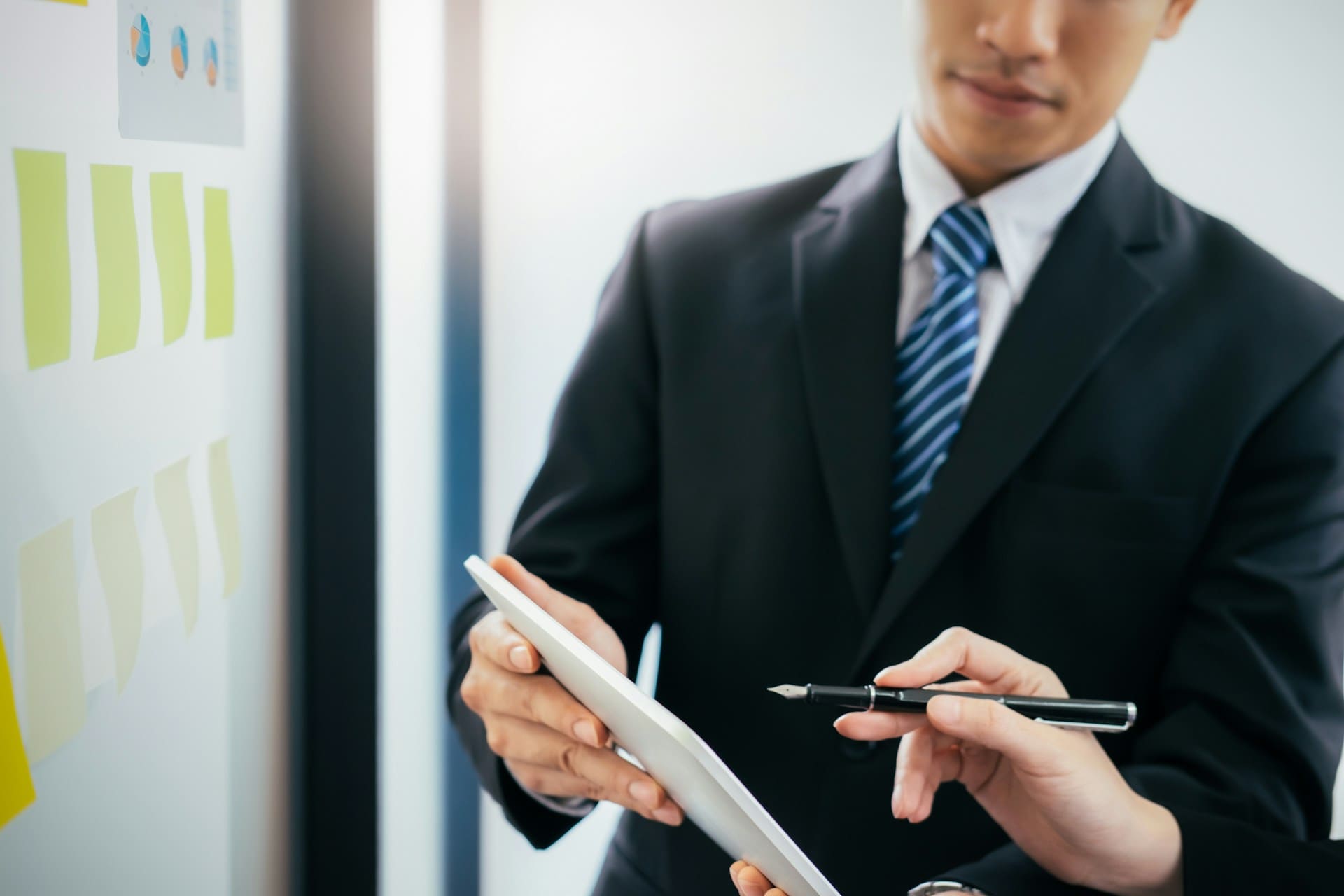 Two people in business attire discuss operational efficiency while reviewing data on a digital tablet. One gestures with a pen, the other holds the device, as sticky notes on a whiteboard suggest an active business consulting session. - Market Design Team: Define. Structure. Expand.